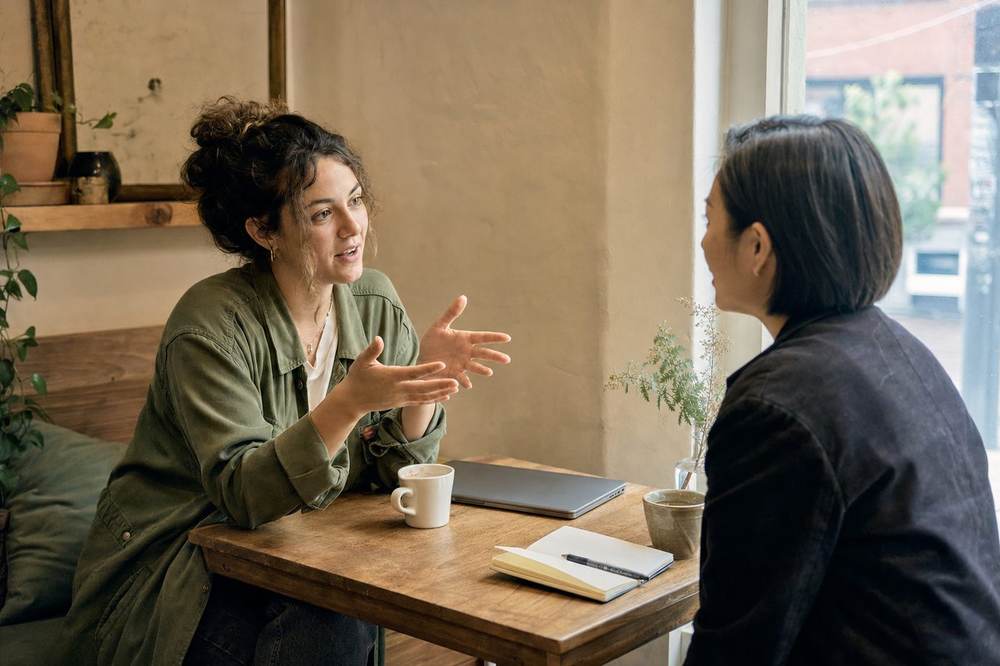 Two people talking across a small wooden table with a notebook and coffee.